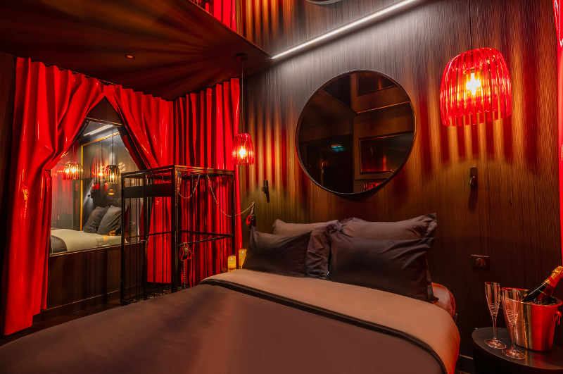 Die Zimmer im Love Hotel Paris besitzen nicht nur Spiegel über dem Bett, sondern auch Fenster zu den benachbarten Zimmern für eine private Peep Show. Das Bild zeigt ein SM-Zimmer mit Käfig und geöffnetem Vorhang mit Blick auf das Bett nebenan